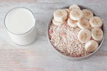 Bowl of dry oat flakes oatmeal with dry berries and sliced banana and glass of wilk on wooden table.
