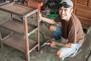 Asian welder smiles at the camera holding an iron rack while welding using an electric welder in a welding workshop