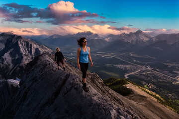 Fototapeta premium Adventurous Girl is hiking up a rocky mountain. Dramatic Colorful Sunset Sky. Taken from Mt Lady MacDonald, Canmore, Alberta, Canada. Epic Adventure Travel