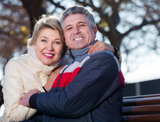 Mature husband and wife hugging each other sitting on park bench