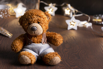 Furry little teddy bear toy with brown fur wearing light blue pants sitting on dark wooden table against background of defocus lights of christmas or new year garland decoration. Image with copy space