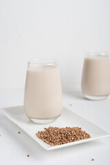 Buckwheat in form of seeds and glass of gluten-free milk which is source of protein, amino acids, manganese and vitamins for healthy dieting on rectangular plate on white wooden background. Vertical