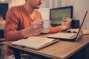 A man hold cup of coffee and looking screen laptop in workplace.