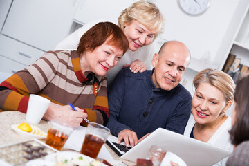 Mature women and their friend are looking at notebook and talking at home.