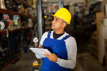 Warehouse male worker in protective helmet and overalls counts the quantity of goods and writes in...