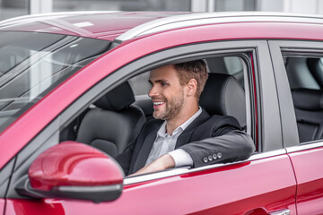 Smiling young male sitting in red car, elbow out of window