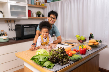 Young Asian man in black frame eyeglasses and lovely little African girl standing behind wooden kitchen table with various vegetables and fruits, looking and smiling together at camera.