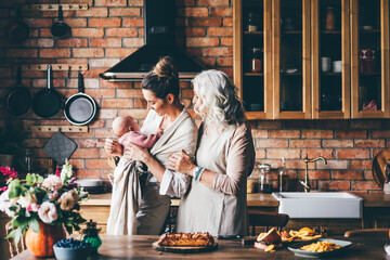 Brunette with holds baby and senior lady looking to the granddaughter preparing dinner in kitchen.