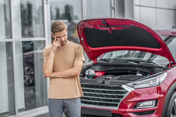 Upset male driver standing in front of broken car with open hood