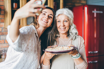 joyful brunette and grey haired mother make selfie with raw pumpkin pie on plate and smile in...