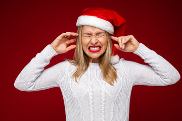 Studio photo of stressed young lady closing her eyes and ears and cringing while wearing Santa hat