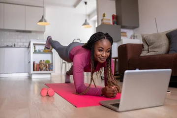 Foto op Plexiglas Young african america woman at home doing exercise in front of open laptop, repeating instructions by professional online fitness trainer. © Zoran Zeremski