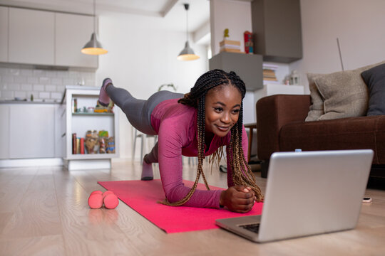 Young african america woman at home doing exercise in front of open laptop, repeating instructions by professional online fitness trainer.