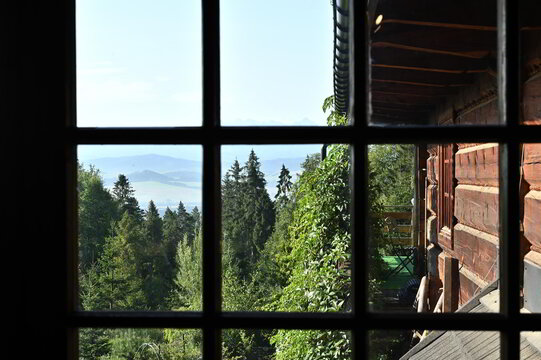 Beautiful view on green trees forest, blue sky and mountains through a wooden window frame of an old house cottage on a slope of a hill in southern Poland