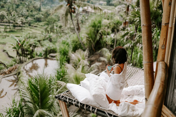 Young travel blogger and influencer girl relaxing in a bamboo house hotel in Bali surrounded by...