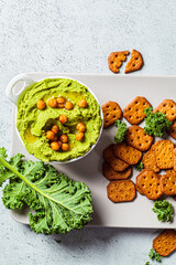 Green kale hummus in white bowl with crackers. Vegan chickpea dip, top view. Healthy vegetarian food concept.