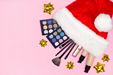 Eye shadow palette kit, lipstick, foundation, make-up brushes in red Santa hat with golden gift bows on pink background. Christmas makeup concept with copy space