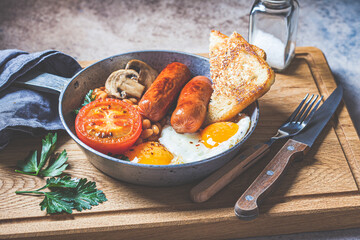 English breakfast with sausages, beans, fried eggs, toast, mushrooms and tomato in frying pan, dark background. English breakfast concept.