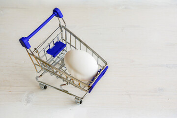 Single white chicken egg in small shopping trolley. Mini cart with egg on wooden white background.