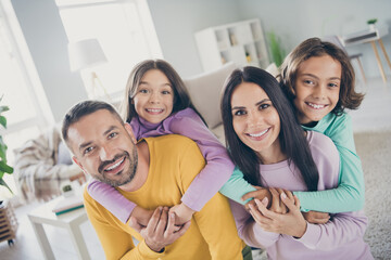 Photo of full family four people parents hold piggy back kids wear colorful sweater in living room indoors