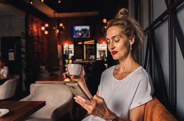Smiling senior woman sitting in coffee shop drinking coffee and using smartphone.