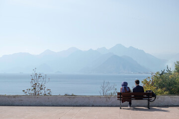 Two lovers are sitting on a bench on the embankment by the ocean against the background of mountains in the haze
