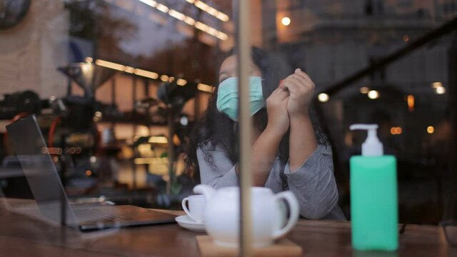 Young African Woman Sitting At Local Cafe In Medical Mask And Using Antiseptic For Hands. Pretty Female Sitting At Table With Personal Wireless Laptop.