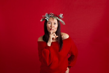 close up portrait of an Asian young woman in a Christmas wreath with a finger at her lips and asking for silence, on a red background.