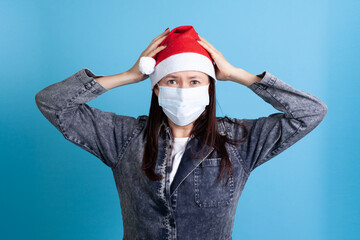 terrified Asian young woman wearing a Santa Claus hat and a medical protective mask on her face, holding her head with her hands.