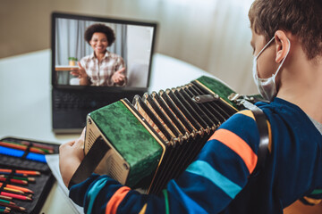 Focused boy playing accordion guitar and watching online course on laptop while practicing at home....