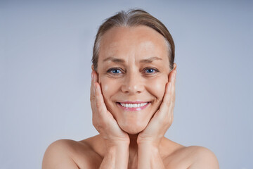 Portrait of a smiling mature woman with her hands to her cheeks, looking at the camera against a gray background. Anti-wrinkle facial skin care concept.