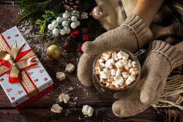 The female hands mittens Christmas gifts and hot cocoa with marshmallows on the wooden table.