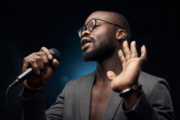 portrait photo of a dark-skinned handsome guy in grey jacket and glasses  on a dark blue background, singing in a microphone  and waving his hand