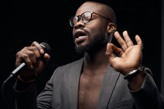 Portrait Of A Dark-skinned Handsome Guy In Grey Jacket And Glasses  On A Black Background, Singing In A Microphone In His Hand With Closed Eyes