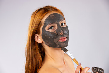  A young red-haired girl applies a clay mask to her face with a brush. Photo over white background with empty side space.