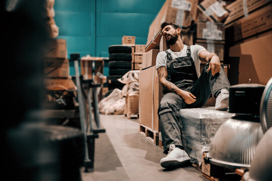 Lazy bearded tattooed worker sleeping on the boxes in working hours. Storage interior.