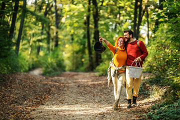 Obraz premium Young happy couple in love hugging and walking in nature. Couple is holding picnic equipment. Woman is pointing at something.