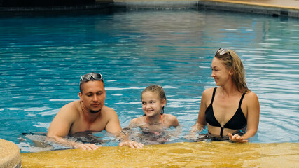 The mother and father with little daughter have fun in the pool. Mom and dad plays with the child. The family enjoy summer vacation in a swimming.