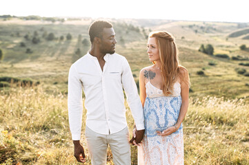 Multiethnic married couple waiting for a baby. A white pregnant woman and a black man walking holding hands on the meadow