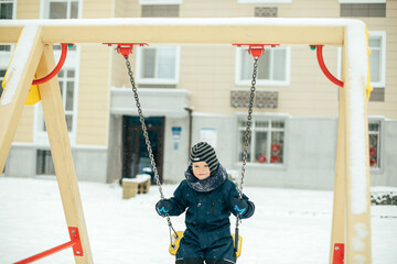 Little boy in winter cloth swinging on a swing in snowfall. Child playing outdoors on playground