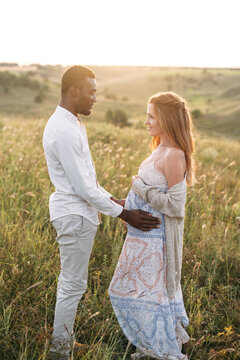 Relaxed multiracial pregnant couple in the park outdoor. An african american husband gently touching tummy of his charming pregnant wife