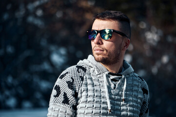 Young confident bearded man brutally posing in a winter forest in sunglasses and a knitted sweater