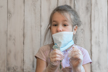 Little girl with medical mask and disposable gloves checks gloves for integrity, anti-coronavirus in pink t-shirt, wooden background photos