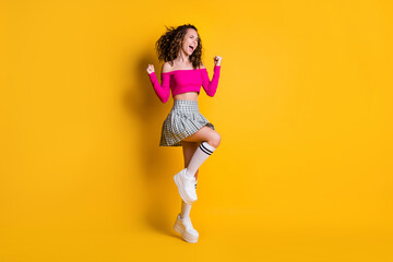 Fototapeta na wymiar Photo portrait of girl celebrating victory standing on one leg fists up wearing casual pink crop-top checkered skirt long socks white sneakers isolated on vivid yellow colored background