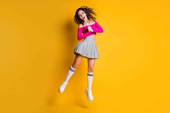 Photo portrait of curly brunette girl jumping holding hands together screaming wearing casual fuchsia crop-top plaid skirt knee-high socks white shoes isolated on vivid yellow colored background