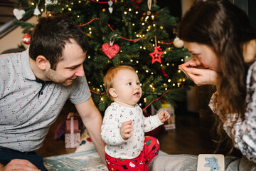 Toddler boy in a Christmas costume and mom, dad sitting on the floor in room. The child with family plays with toys near the Christmas tree. Happy winter holidays. Happy New Year and Merry Christmas.