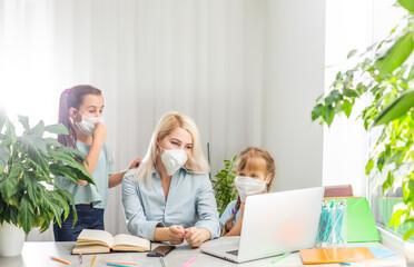 Young woman in medical mask pointing at data on laptop for little girl while sitting at table and working on homework assignment together