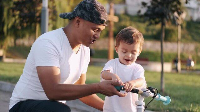 Little kid is learning to ride a bike with his dad outdoors in a pubic park. Family time, summer activities.