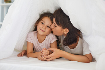 Loving young mother kisses her little daughter on the bed under the blanket in the bedroom.