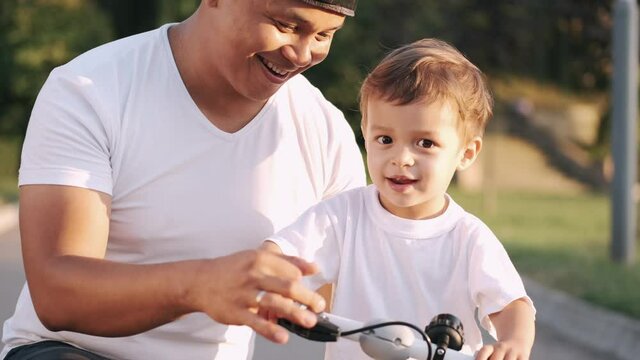 Little kid is learning to ride a bike with his dad outdoors in a pubic park. Family time, summer activities.